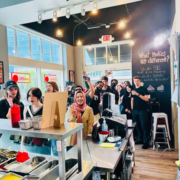 Diverse group of young people smiling and posing behind a restaurant counter with food prep stations.