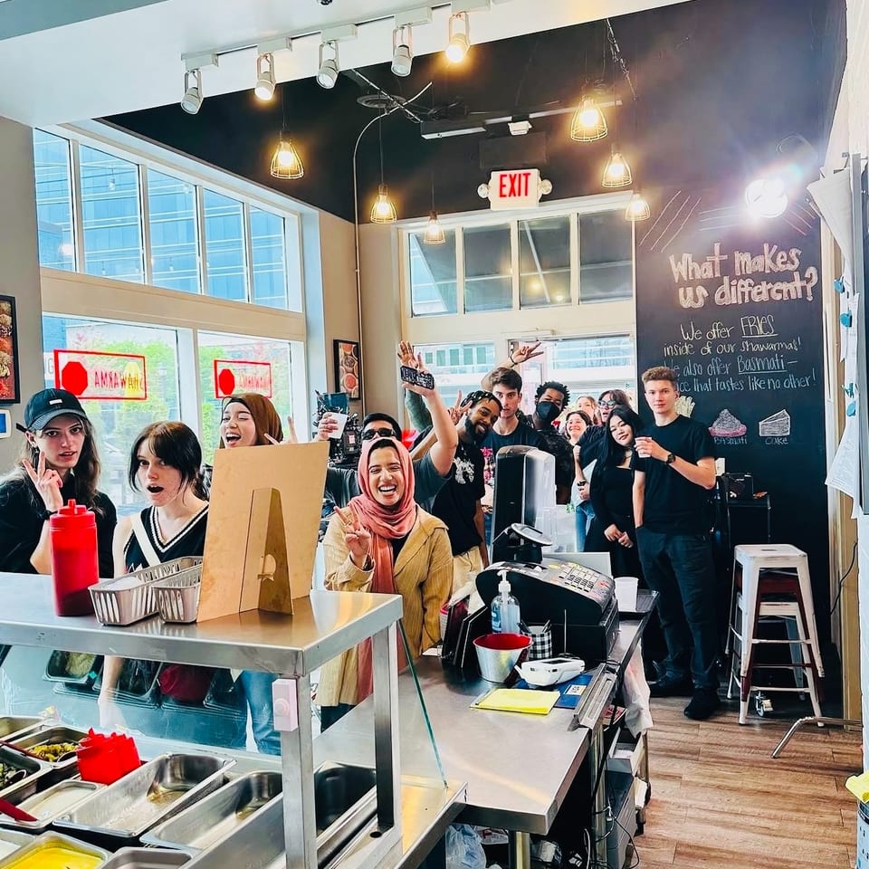 Diverse group of young people smiling and posing behind a restaurant counter with food prep stations.