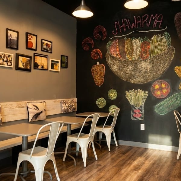 Restaurant interior with a chalkboard mural of shawarma ingredients and framed pictures on the opposite wall.