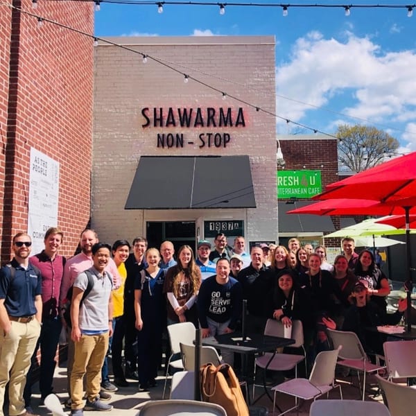 A large group of smiling people gathered outside Shawarma Non-Stop restaurant on a sunny day.