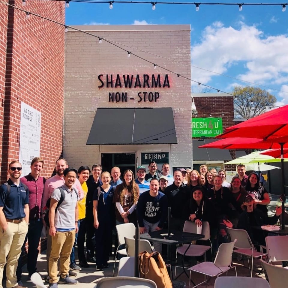A large group of smiling people gathered outside Shawarma Non-Stop restaurant on a sunny day.
