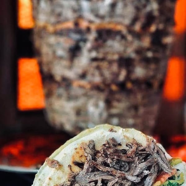 A hand holds a shawarma wrap with meat, lettuce, tomato, and sauce, in front of a meat spit.